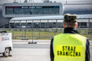 Funkcjonariusz w żółtej kamizelce z napisem Straż Graniczna stoi na płycie lotniska. W tle budynek terminala. Funkcjonariusz w żółtej kamizelce z napisem Straż Graniczna stoi na płycie lotniska. W tle budynek terminala.