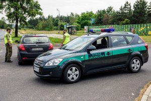 Na pierwszym planie na drodze stoi oznakowany samochód Straży Granicznej. Za nim funkcjonariusz kontrolują zatrzymany pojazd. Na pierwszym planie na drodze stoi oznakowany samochód Straży Granicznej. Za nim funkcjonariusz kontrolują zatrzymany pojazd.