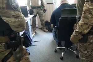 Zatrzymany siedzi przy biurku, obok stoją funkcjonariusze WZD Zatrzymany siedzi przy biurku, obok stoją funkcjonariusze WZD