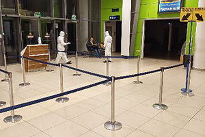 Funkcjonariusze SG ubrani w kombinezony ochronne stoją w budynku terminala. Na drugim planie siedzi zatrzymany cudzoziemiec. Funkcjonariusze SG ubrani w kombinezony ochronne stoją w budynku terminala. Na drugim planie siedzi zatrzymany cudzoziemiec.