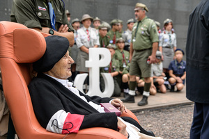 Uczestniczka powstania warszawskiego siedzi na fotelu. W tle harcerze trzymający symbol Polski Walczącej. Uczestniczka powstania warszawskiego siedzi na fotelu. W tle harcerze trzymający symbol Polski Walczącej.