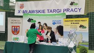 Stoisko Powiatowego Urzędu Pracy w Mławie. U góry wisi baner z napisem dziesiąte mławskie targi pracy. Stoisko Powiatowego Urzędu Pracy w Mławie. U góry wisi baner z napisem dziesiąte mławskie targi pracy.