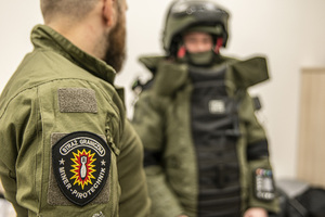 Na pierwszym planie naszywka na ramieniu pirotechnika. Na drugim planie stoi pirotechnik ubrany w kombinezon. Na pierwszym planie naszywka na ramieniu pirotechnika. Na drugim planie stoi pirotechnik ubrany w kombinezon.