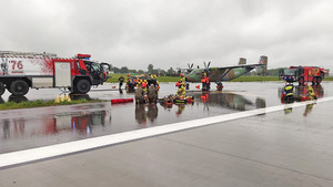 Na płycie lotniska stoją wozy strażackie oraz samolot. Służby udzielają pomocy poszkodowanym. Na płycie lotniska stoją wozy strażackie oraz samolot. Służby udzielają pomocy poszkodowanym.
