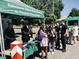 Funkcjonariusze prezentują sprzęt służbowy na stoisku promocyjnym. Przed namiotem stoi młodzież. Funkcjonariusze prezentują sprzęt służbowy na stoisku promocyjnym. Przed namiotem stoi młodzież.
