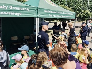 Duża grupa zwiedzających stoi przed namiotami. Po prawej stronie widać funkcjonariuszy, Chłopiec celuje z broni. Duża grupa zwiedzających stoi przed namiotami. Po prawej stronie widać funkcjonariuszy, Chłopiec celuje z broni.