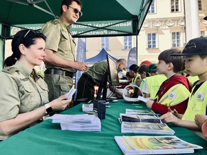 Na stoliku leżą ulotki promocyjne. Po lewej stronie funkcjonariusze Straży Granicznej, na przeciwko nich stoją dzieci w żółtych kamizelkach. Na stoliku leżą ulotki promocyjne. Po lewej stronie funkcjonariusze Straży Granicznej, na przeciwko nich stoją dzieci w żółtych kamizelkach.