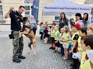 Pies w wyskoku łapie zabawkę, którą trzyma przewodnik. Po prawej stronie stoją uśmiechnięte dzieci i opiekunki. Pies w wyskoku łapie zabawkę, którą trzyma przewodnik. Po prawej stronie stoją uśmiechnięte dzieci i opiekunki.
