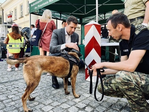 Wojewoda Mazowiecki Pan Tobiasz Bocheński i funkcjonariusz kucają przy psie służbowym. Na drugim planie stoi słup graniczny i namiot. Wojewoda Mazowiecki Pan Tobiasz Bocheński i funkcjonariusz kucają przy psie służbowym. Na drugim planie stoi słup graniczny i namiot.