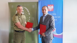 Dyrektor IAS w Warszawie Wojciech Kustra i Komendant NwOSG ściskają sobie dłonie. W lewych rękach trzymają czerwone teczki. W tle widać loga instytucji. Dyrektor IAS w Warszawie Wojciech Kustra i Komendant NwOSG ściskają sobie dłonie. W lewych rękach trzymają czerwone teczki. W tle widać loga instytucji.