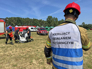 Na pierwszym planie kierujący działaniami ratowniczymi. Na drugim planie strażacy rozstawiają sprzęt. W tle czerwone pojazdy. Na pierwszym planie kierujący działaniami ratowniczymi. Na drugim planie strażacy rozstawiają sprzęt. W tle czerwone pojazdy.
