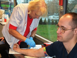Mężczyzna w okularach siedzi z zaciśniętą dłonią. Kobieta w białym fartuchu pobiera krew. Mężczyzna w okularach siedzi z zaciśniętą dłonią. Kobieta w białym fartuchu pobiera krew.
