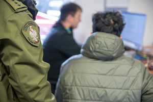 Po lewej widoczna naszywka na rękawie kurtki służbowej. Po prawej funkcjonariusz wpisuje dane do komputera. Tyłem siedzi cudzoziemiec. Po lewej widoczna naszywka na rękawie kurtki służbowej. Po prawej funkcjonariusz wpisuje dane do komputera. Tyłem siedzi cudzoziemiec.