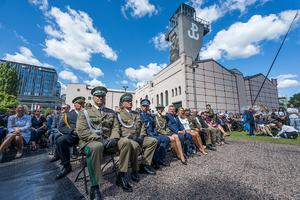 Przedstawiciele służb i goście siedzą na krzesłach. W tle budynek Muzeum Powstania Warszawskiego. Przedstawiciele służb i goście siedzą na krzesłach. W tle budynek Muzeum Powstania Warszawskiego.