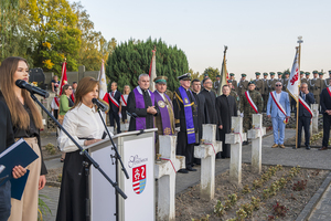 Dyrektor szkoły Pani Marta Bińkowska stoi przy mównicy, na drugim planie za krzyżami stoją księża, poczty sztandarowe oraz kompania. Dyrektor szkoły Pani Marta Bińkowska stoi przy mównicy, na drugim planie za krzyżami stoją księża, poczty sztandarowe oraz kompania.