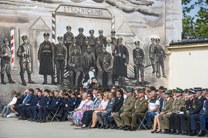 Uczestnicy uroczystości siedzą na krzesłach, na drugim planie widać mural na ścianie. Uczestnicy uroczystości siedzą na krzesłach, na drugim planie widać mural na ścianie.
