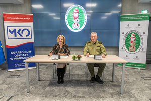 Pani Wioletta Krzyżanowska i płk SG Dariusz Lutyński siedzą za stołem, za nimi po bokach stoją rollupy z logo instytucji. Pani Wioletta Krzyżanowska i płk SG Dariusz Lutyński siedzą za stołem, za nimi po bokach stoją rollupy z logo instytucji.