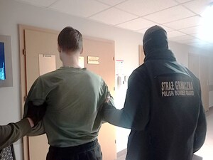 Zatrzymany mężczyzna stoi przed drzwiami, funkcjonariusz trzyma go za rękę. Zatrzymany mężczyzna stoi przed drzwiami, funkcjonariusz trzyma go za rękę.