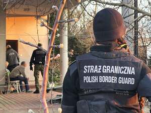 W głębi funkcjonariusze stoją przed drzwiami domu, po prawej stronie funkcjonariuszka w kamizelce z napisem Straż Graniczna. W głębi funkcjonariusze stoją przed drzwiami domu, po prawej stronie funkcjonariuszka w kamizelce z napisem Straż Graniczna.