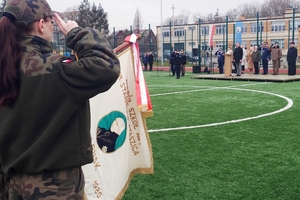 Po lewej stronie kadetka oddaje honor, widoczny jest sztandar. W głębi trybuna, na której stoją goście. Po lewej stronie kadetka oddaje honor, widoczny jest sztandar. W głębi trybuna, na której stoją goście.