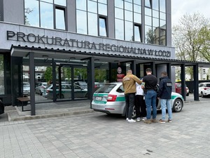 Funkcjonariusze wprowadzają zatrzymanego do samochodu. Na drugim planie budynek prokuratury. Funkcjonariusze wprowadzają zatrzymanego do samochodu. Na drugim planie budynek prokuratury.