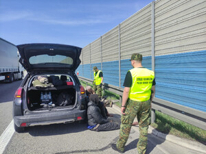 Przy zatrzymanym samochodzie siedzą cudzoziemcy, obok nich w żółtych kamizelkach stoją funkcjonariusze Straży Granicznej. Przy zatrzymanym samochodzie siedzą cudzoziemcy, obok nich w żółtych kamizelkach stoją funkcjonariusze Straży Granicznej.