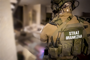 Na pierwszym planie stoi funkcjonariusz Straży Granicznej, w głębi rozmyte wnętrze mieszkania. Na pierwszym planie stoi funkcjonariusz Straży Granicznej, w tle rozmyte wnętrze mieszkania.