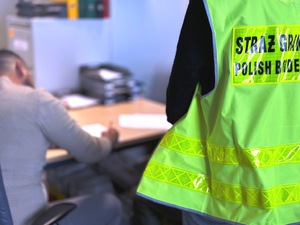 Cudzoziemiec podpisuje dokumenty, obok stoi funkcjonariuszka w żółtej kamizelce. Cudzoziemiec podpisuje dokumenty, obok stoi funkcjonariuszka w żółtej kamizelce.