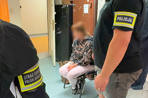 Zatrzymana siedzi na krześle obok stoją funkcjonariusze. Zatrzymana siedzi na krześle obok stoją funkcjonariusze.